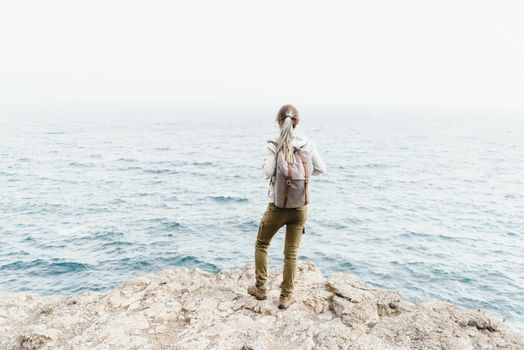 Traveler girl with backpack standing on rocky coastline and looking at sea.