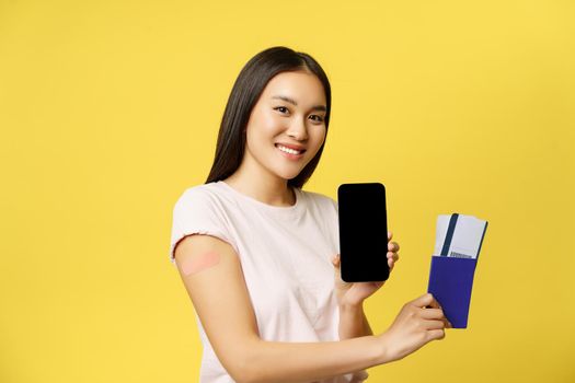 Smiling asian girl vaccinated, showing passport tickets and smartphone screen, app interface, standing over yellow background.