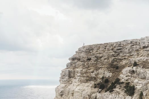 Small figure of female traveler standing on edge of cliff over sea on background of cloudy sky. Concept of minimal person in massive landscape.