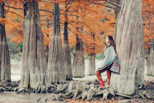 Beautiful boho style young woman walking in autumn park among cypresses.