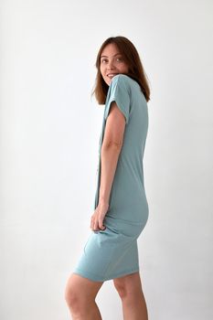 Side view of positive slender female wearing blue dress in casual style standing on white background in studio and looking away