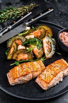 Fish meal with Roasted Salmon Fillet Steaks and arugula tomato salad on a plate. Black background. Top view.