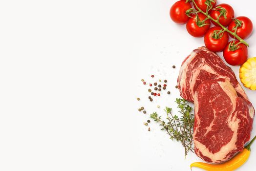 two fresh marbled beef steaks on a white plate with vegetables cherry tomatoes on a sprig, corn, spicy yellow pepper, a sprig of rosemary, salt and different varieties of peppers