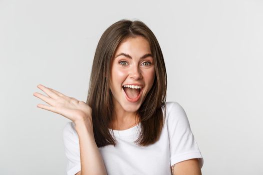 Close-up of excited beautiful brunette girl looking amused, waving hand to say hello, white background.