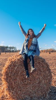 A girl of European appearance in a gray coat stands in a field near a larger bale with hay against a blue sky