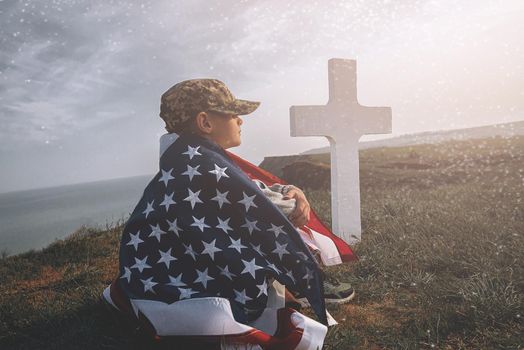 child teenager wrapped in an American flag sits on the grass near the grave of his father, a soldier hero of America