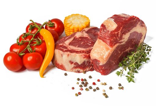 two fresh marbled beef steaks on a white plate with vegetables cherry tomatoes on a sprig, corn, spicy yellow pepper, a sprig of rosemary, salt and different varieties of peppers