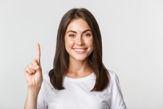 Close-up of cheerful attractive young woman smiling, showing one finger, white background.