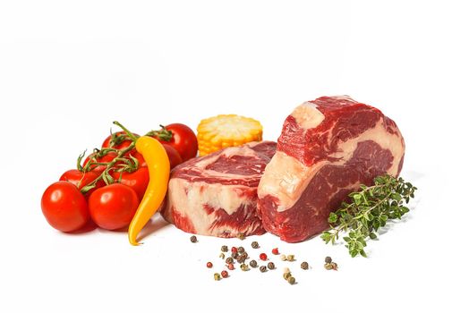 two fresh marbled beef steaks on a white plate with vegetables cherry tomatoes on a sprig, corn, spicy yellow pepper, a sprig of rosemary, salt and different varieties of peppers