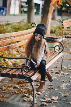 Stylish baby girl 4-5 year old wearing knitted hat, sunglasses, boots, fur coat sitting on the bench in park. Looking at camera. Autumn fall season