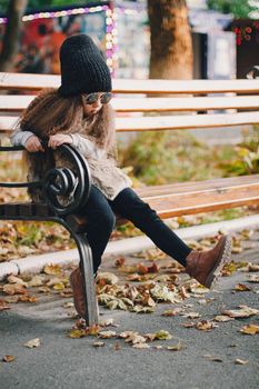 Stylish baby girl 4-5 year old wearing knitted hat, sunglasses, boots, fur coat sitting on the bench in park. Looking at camera. Autumn fall season