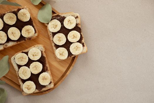 Three banana white bread toasts smeared with chocolate butter that lie on a cutting board with a sprig of leaves on craft paper background. top view with area for text