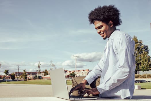 Young African American Guy Working On Laptop Outside, Sitting On Bench In Park. Side View