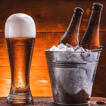 two bottles of beer in a bucket with ice and a glass of beer with lush foam next to a dark background