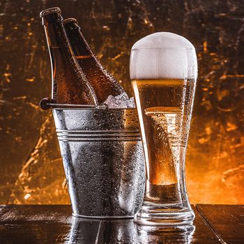 two bottles of beer in a bucket with ice and a glass of beer with lush foam next to a dark background