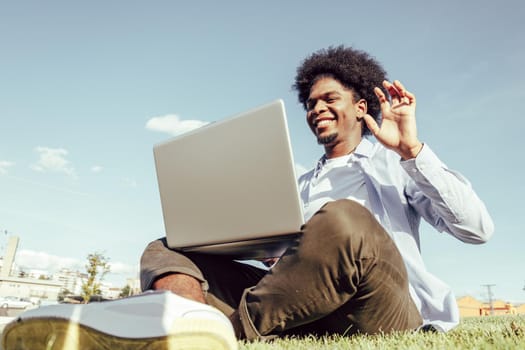 Black ethnicity man with afro hair using his laptop sitting on the grass of an urban park.