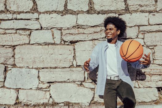 Happy afro hair dark skinned man with basket ball leaning on wall. Copy space