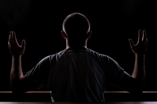 Silhouette of a young guy who sits in church and praying with hands raised up. Close-up view from behind. Backlight.