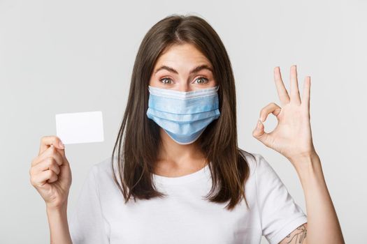 Covid-19, health and social distancing concept. Close-up of smiling brunette girl in medical mask, showing credit card and okay gesture.