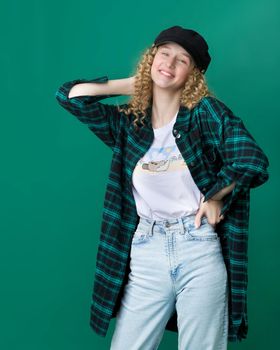 Happy smiling blonde curly girl touching her hair. Portrait of cheerful girl in trendy clothes. Young woman wearing dark plaid cotton shirt, black cap and jeans posing on blue green background