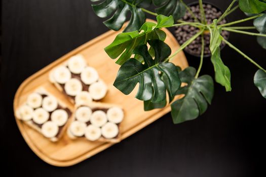 Three banana white bread toasts spread with chocolate butter that lie on a chopping board with a sprig of leaves on a dark background. top view with area for text