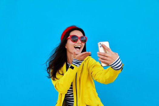 Portrait of young woman taking a selfie by smartphone wearing a knitted white hat on colorful blue background