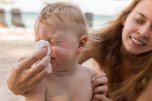 Happy woman keeping handkerchief near nose of sneezing baby while resting on beach on resort