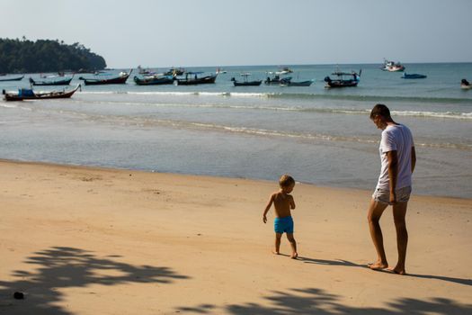 Side view of young man following little boy while walking together on sandy beach in sunny summer day with boats on water in background