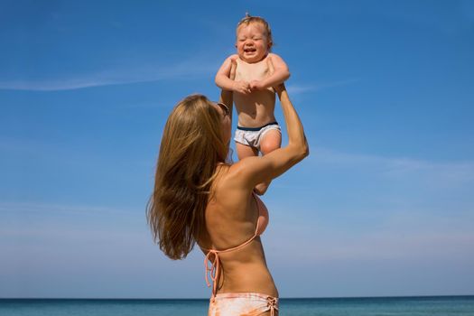 Happy young woman in bikini holding up cute laughing little baby while standing on beach in sunny day with blue sky in background