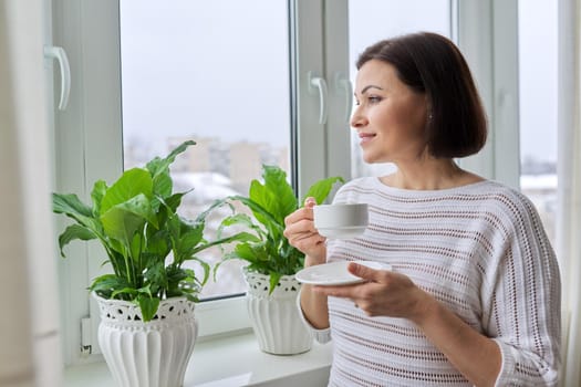 Season winter, snowy day, middle aged smiling woman with cup of coffee looking out the window. Female 40s of age, lifestyle, portrait, copy space