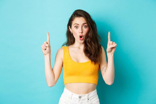 Summer lifestyle concept. Surprised young woman gasping excited, pointing fingers up, showing advertisement with amazed face, standing over blue background.