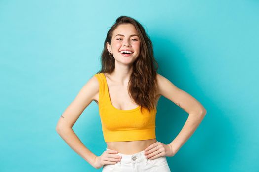 Image of confident and happy stylish woman laughing, holding hands on waist and smiling cheerful, standing carefree in summer outfit, blue background.