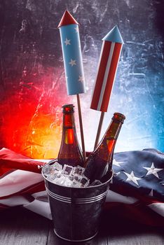 two bottles of beer in an ice bucket with the American flag lying nearby and rockets for fireworks. Independence Day celebration concept