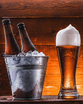 two bottles of beer in a bucket with ice and a glass of beer with lush foam next to a dark background