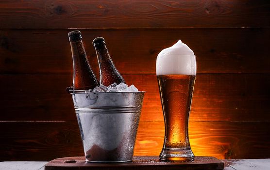 two bottles of beer in a bucket with ice and a glass of beer with lush foam next to a dark background