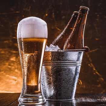 two bottles of beer in a bucket with ice and a glass of beer with lush foam next to a dark background