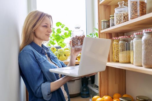 Middle-aged woman in kitchen in pantry with laptop showing cans of food. Female food blogger, online communication, healthy eating video recording, healthy food, technology, hobbies and leisure