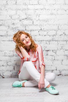Young beautiful caucasian woman student is sitting on the floor near a white brick wall. Girl with red long curly hair with freckles on her face in bright summer clothes.