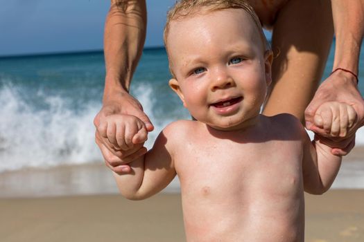 Adorable healthy little undressed baby with wet hair looking at camera while standing with help of crop mother on sandy beach in sunny day