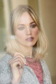 Thoughtful young blonde woman with lost eyes from the window. Beautiful girl at home wearing winter clothes. Pretty female with straight hair and blue eyes.