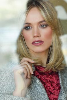 Thoughtful young blonde woman with lost eyes from the window. Beautiful girl at home wearing winter clothes. Pretty female with straight hair and blue eyes.