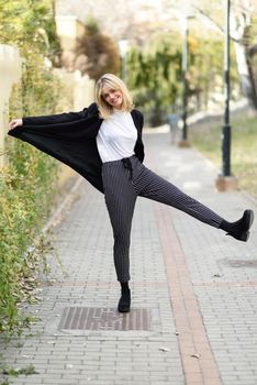 Funny blonde woman smiling in urban background. Young girl wearing black blazer jacket and striped trousers standing in the street. Pretty female with straight hair hairstyle and blue eyes.