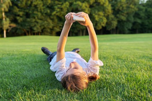 Guy teenager lying on grass with smartphone. Male in white T-shirt with phone in his hands, top view, green lawn background. Youth, technology, lifestyle, adolescence concept