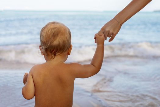 Back view of little child holding hands with crop mother going to water relaxing in sea