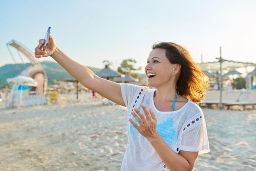 Happy middle-aged woman talking on smartphone using video call, female on sandy sea beach. Summer, vacation, weekend in nature, people of mature age