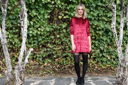 Beautiful blonde woman in green leaves background. Young girl wearing red dress standing in the street. Pretty female with frizzy hairstyle and blue eyes.