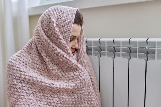 Winter, heating season. A frozen woman in warm woolen sweater under knitted blanket sitting in home room near heating radiator