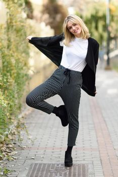 Funny blonde woman smiling in urban background. Young girl wearing black blazer jacket and striped trousers standing in the street. Pretty female with straight hair hairstyle and blue eyes.