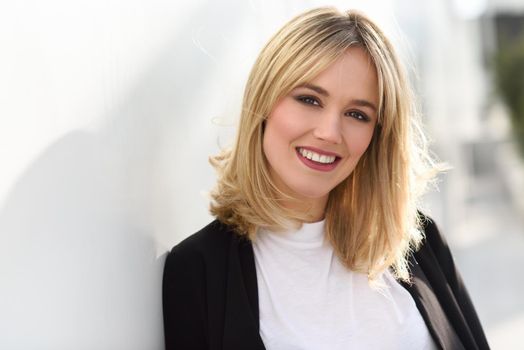 Beautiful blonde woman smiling in urban background. Young girl wearing black blazer jacket standing in the street. Pretty female with straight hair hairstyle and blue eyes.