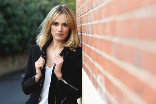 Attractive blonde woman in urban background. Young girl wearing black zipper jacket standing in the street. Pretty female with straight hair hairstyle and blue eyes on brick wall.